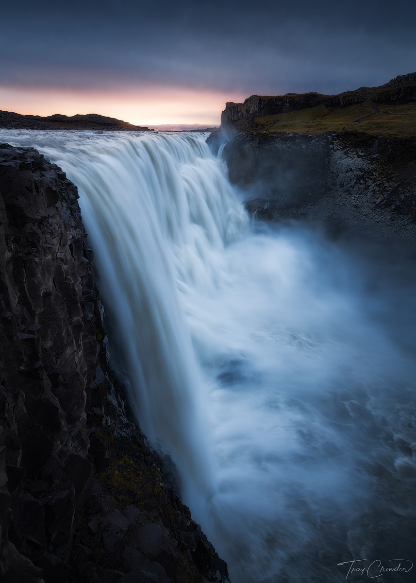 Sunrise light peaks through a blanket of storm clouds on the horizon over Dettifoss waterfall in Iceland.